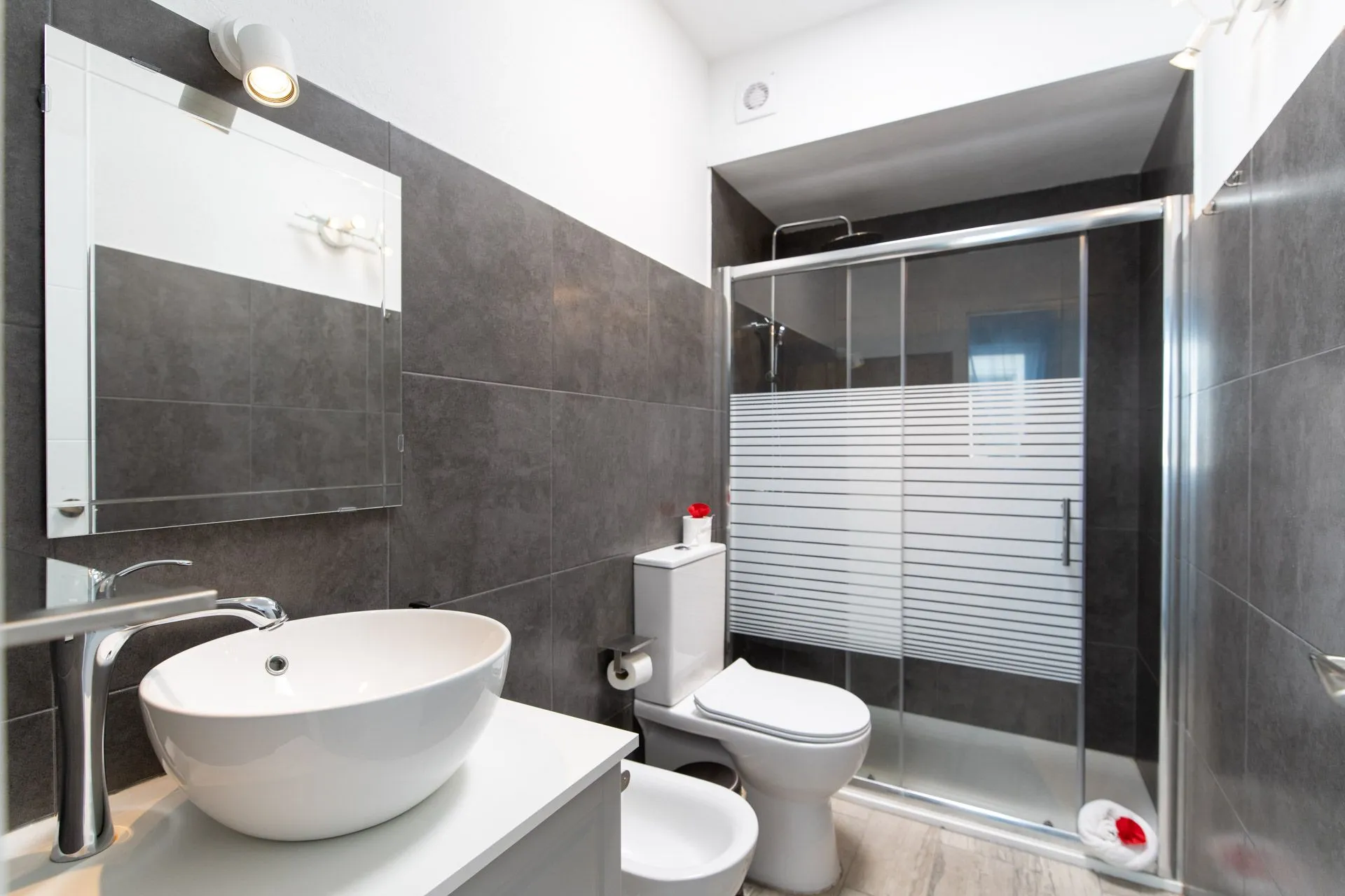 Bagno con piastrelle grigie, doccia in vetro, lavabo e sanitari