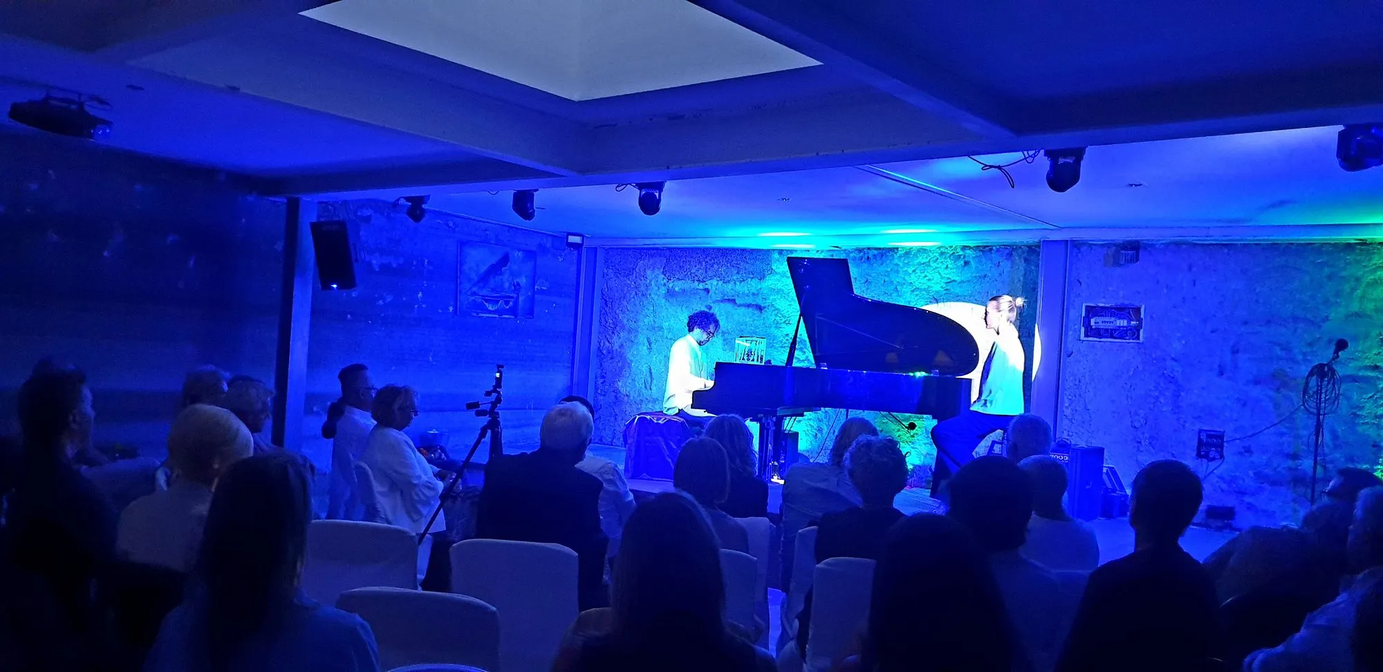 Pianista suona un pianoforte a coda sotto luce blu davanti al pubblico seduto in sala