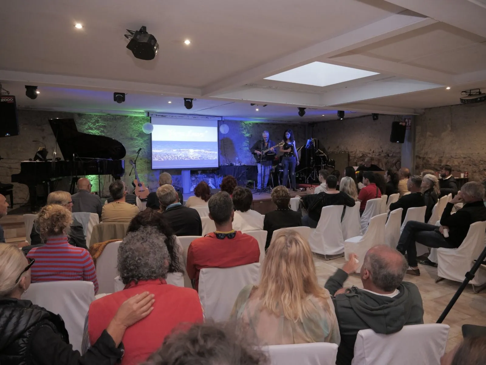 Pubblico seduto assiste a un concerto in sala con musicisti sul palco illuminato